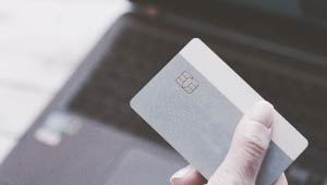 A picture of a hand holding a payment card over a laptop that is blurred in the background.
