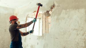 A man with a sledge hammer swinging towards a wall with a large whole in it.